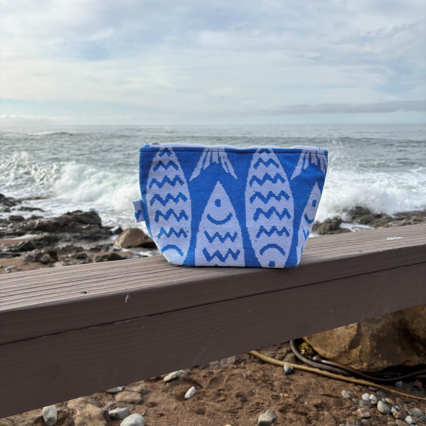 large blue and white fish print makeup bags placed on a wooden bench with a coastal background.