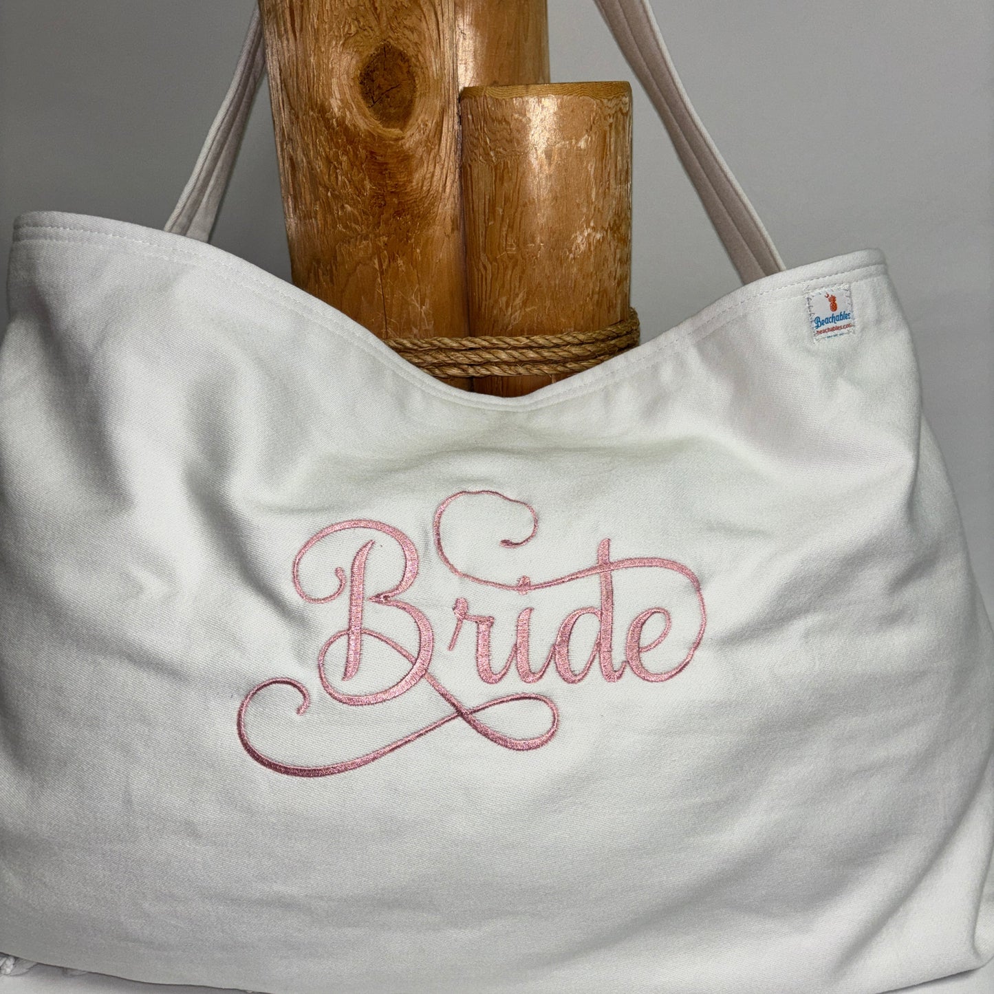 A white canvas tote bag with pink embroidered script reading 'Bride' on the front, shown with wooden cylindrical props against a light gray background.