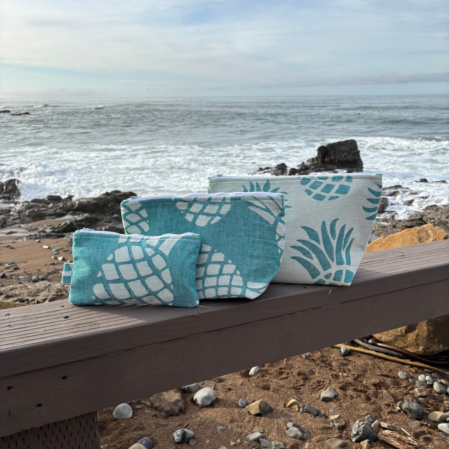 A set of Pineapple makeup bags in various sizes placed on a wooden bench with a coastal background.