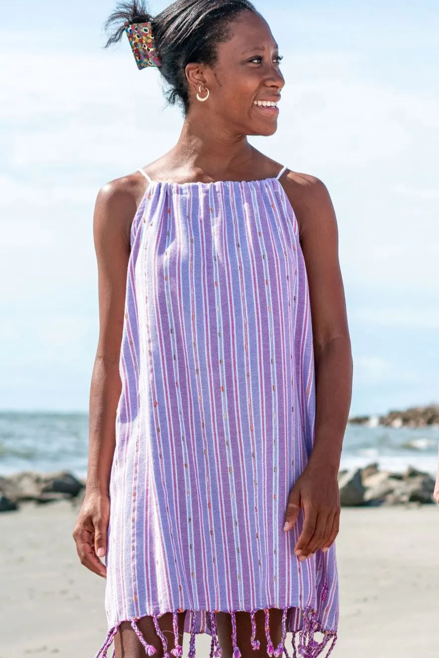 A woman standing on a beach wearing a sleeveless, purple striped mini dress with tassels.