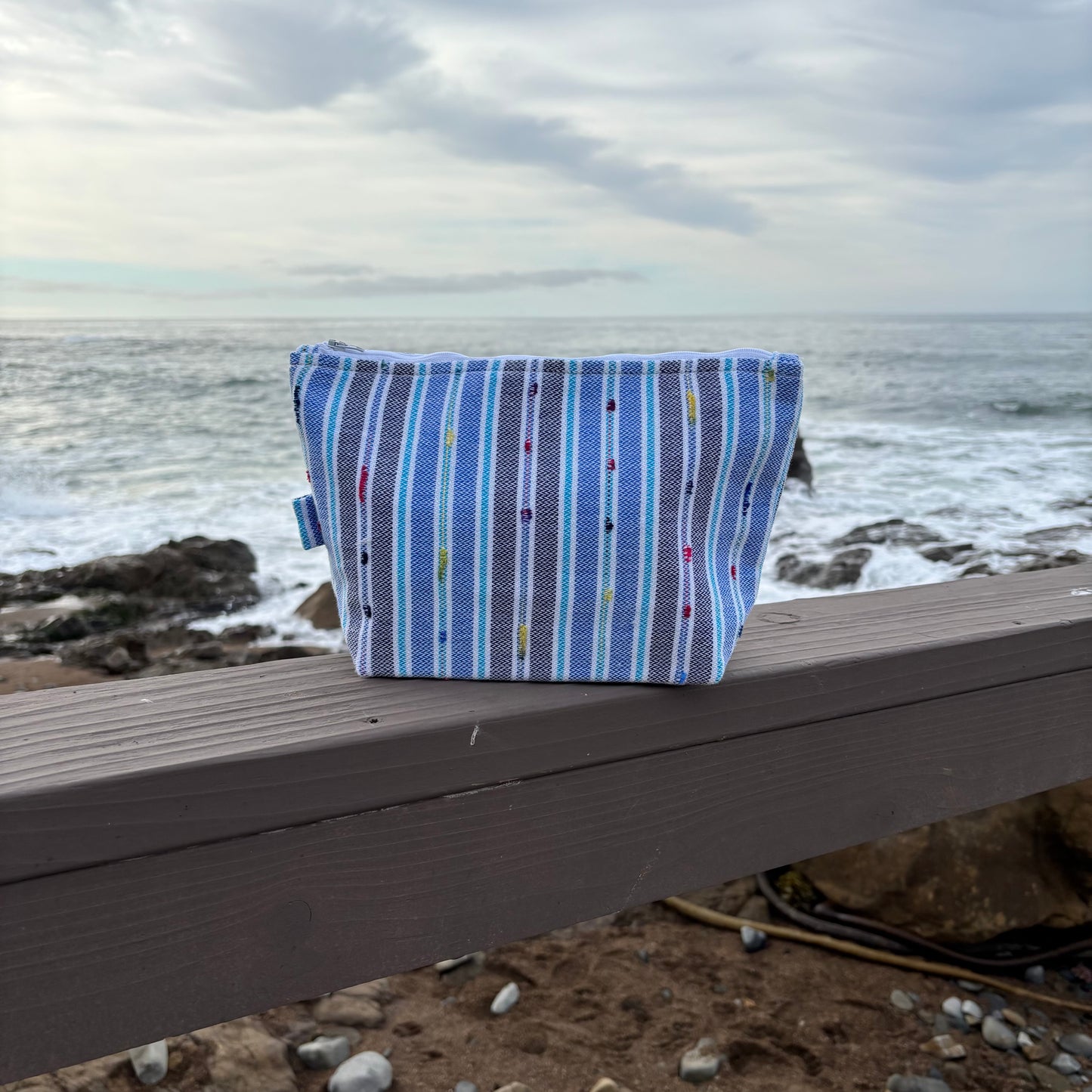 Large sized blue striped makeup bag placed on a surface with a coastal background.
