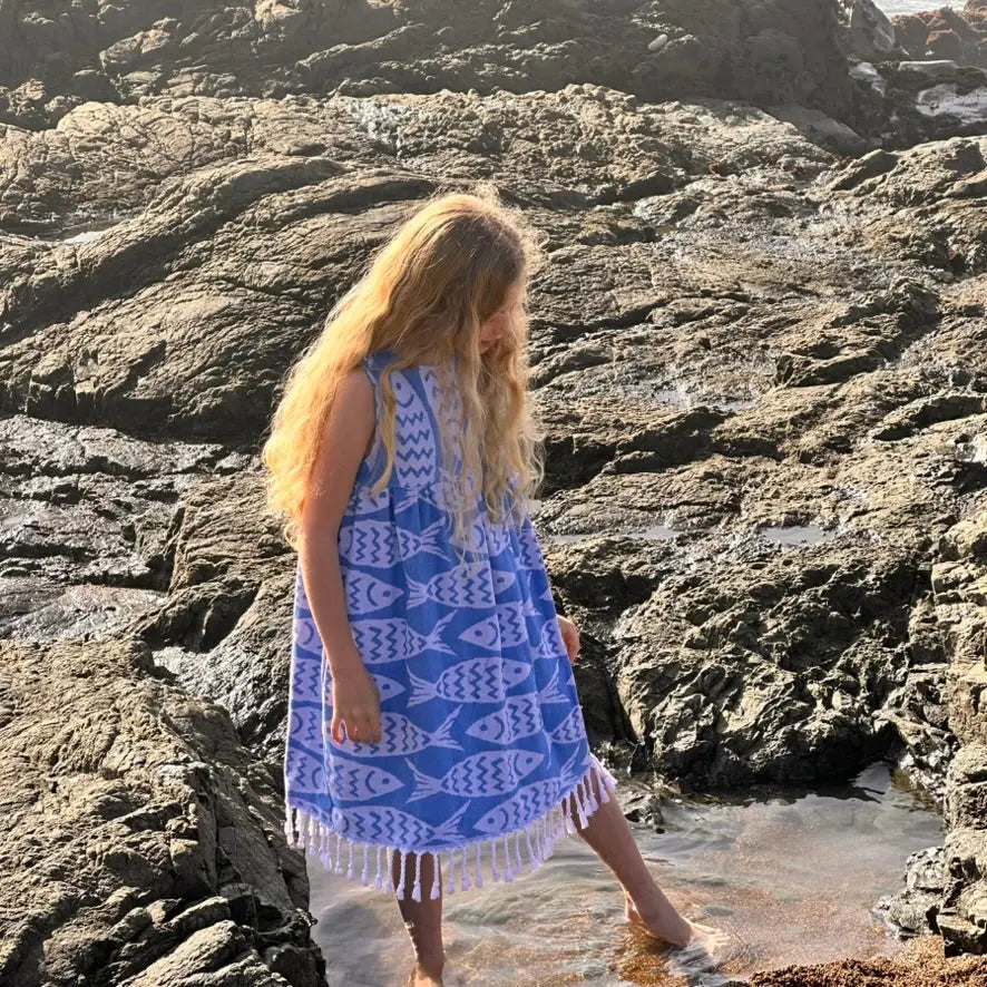 A girl wearing a blue and white fish print sleeveless dress with tassels on a rocky beach