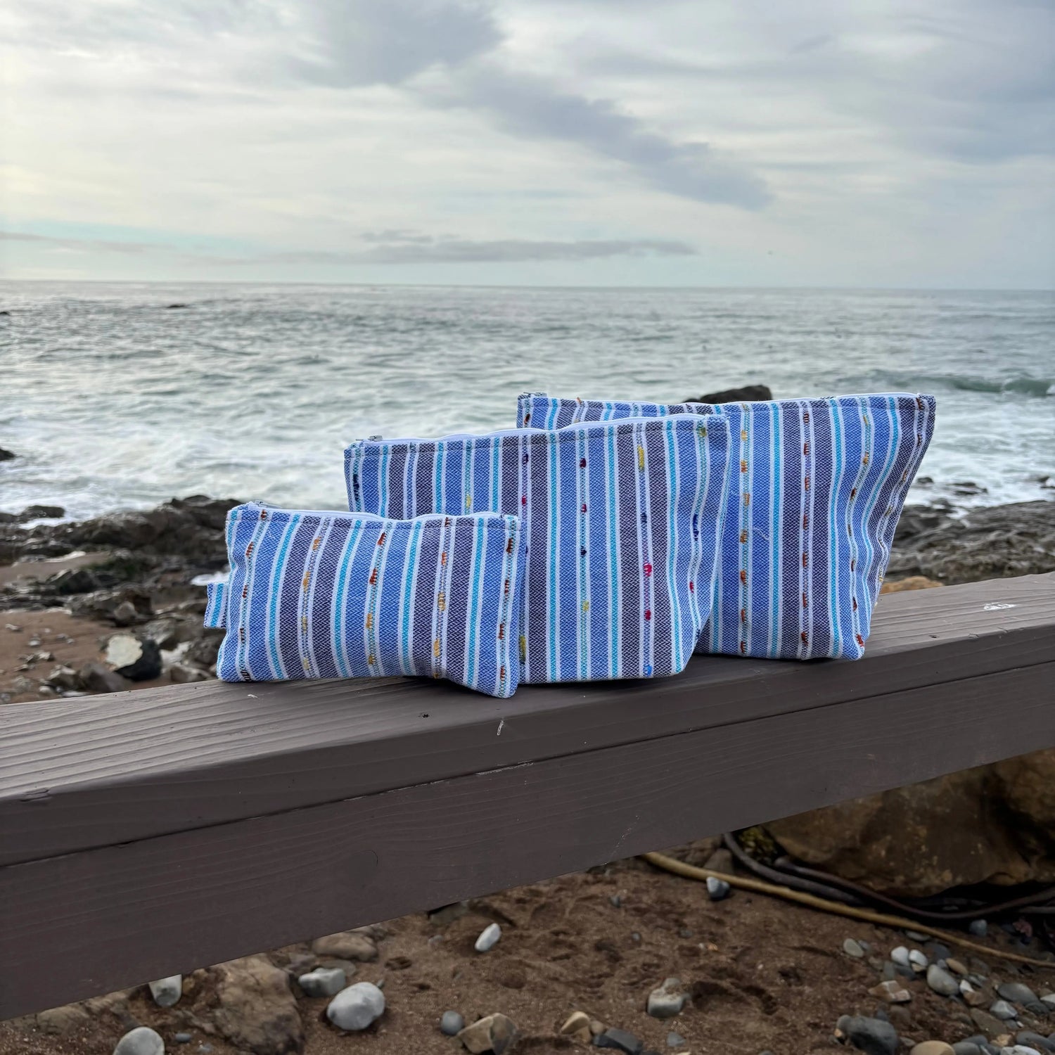 Assortment of different sized blue striped zipper bags displayed on bench with ocean in background