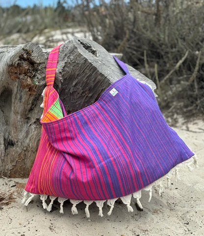 Colorful pink, orange, and purple striped bag with tassels draped over a log on a sandy beach.
