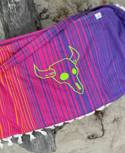 A multicolored striped embroidered bag with a cow skull design, hanging on a wooden structure with a sandy background.