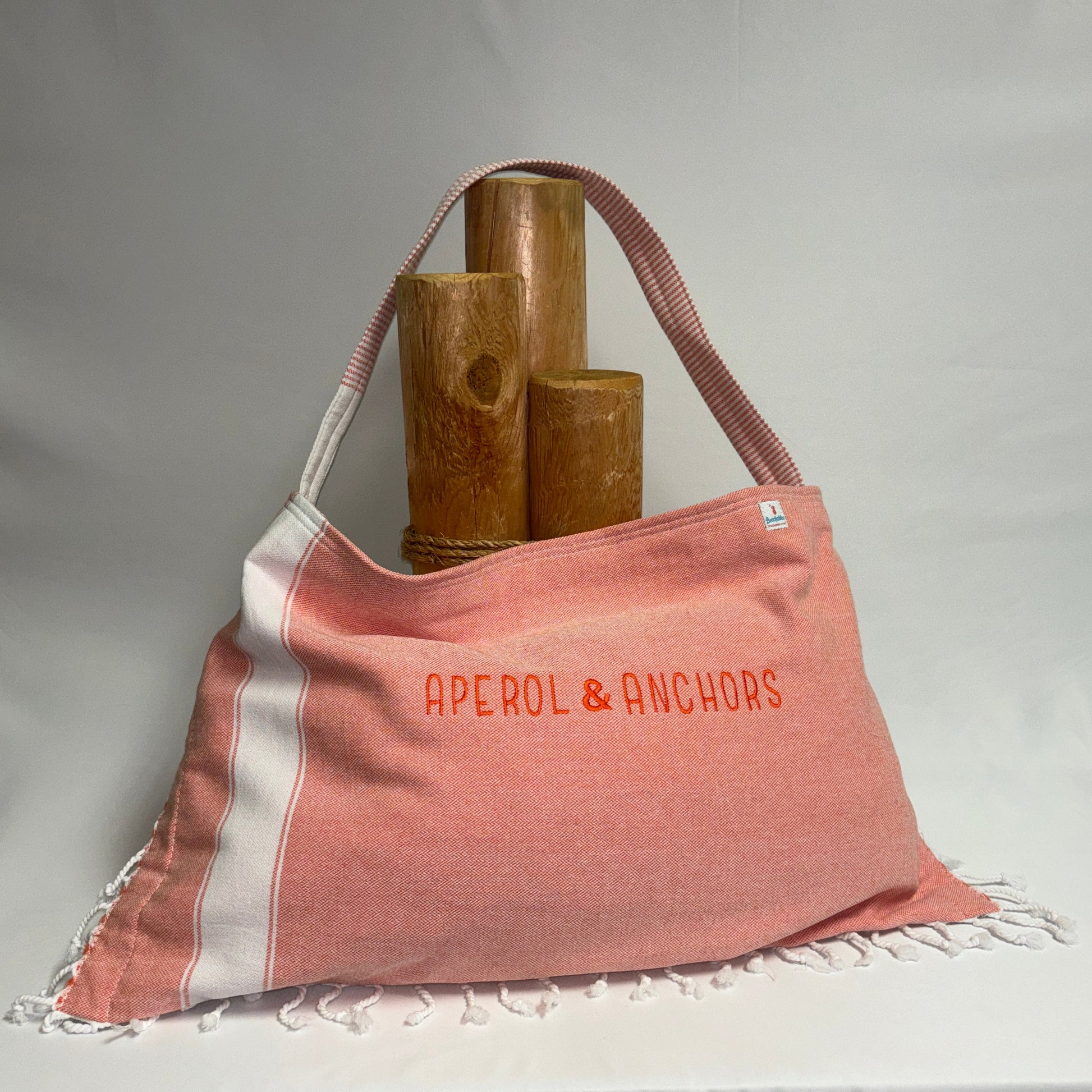 A terracotta pink and white striped Turkish towel tote bag with fringe, featuring the text 'APEROL & ANCHORS' in orange. The bag is styled with three wooden cylinder props against a draped white fabric backdrop.