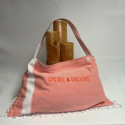 A terracotta pink and white striped Turkish towel tote bag with fringe, featuring the text 'APEROL & ANCHORS' in orange. The bag is styled with three wooden cylinder props against a draped white fabric backdrop.