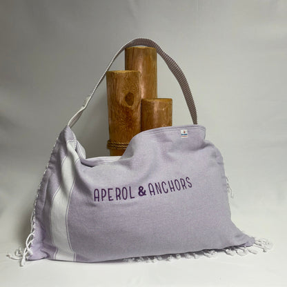A lavender and white fringed shoulder bag with 'APEROL & ANCHORS' printed on the front, displayed on a draped white fabric backdrop with wooden props behind it.