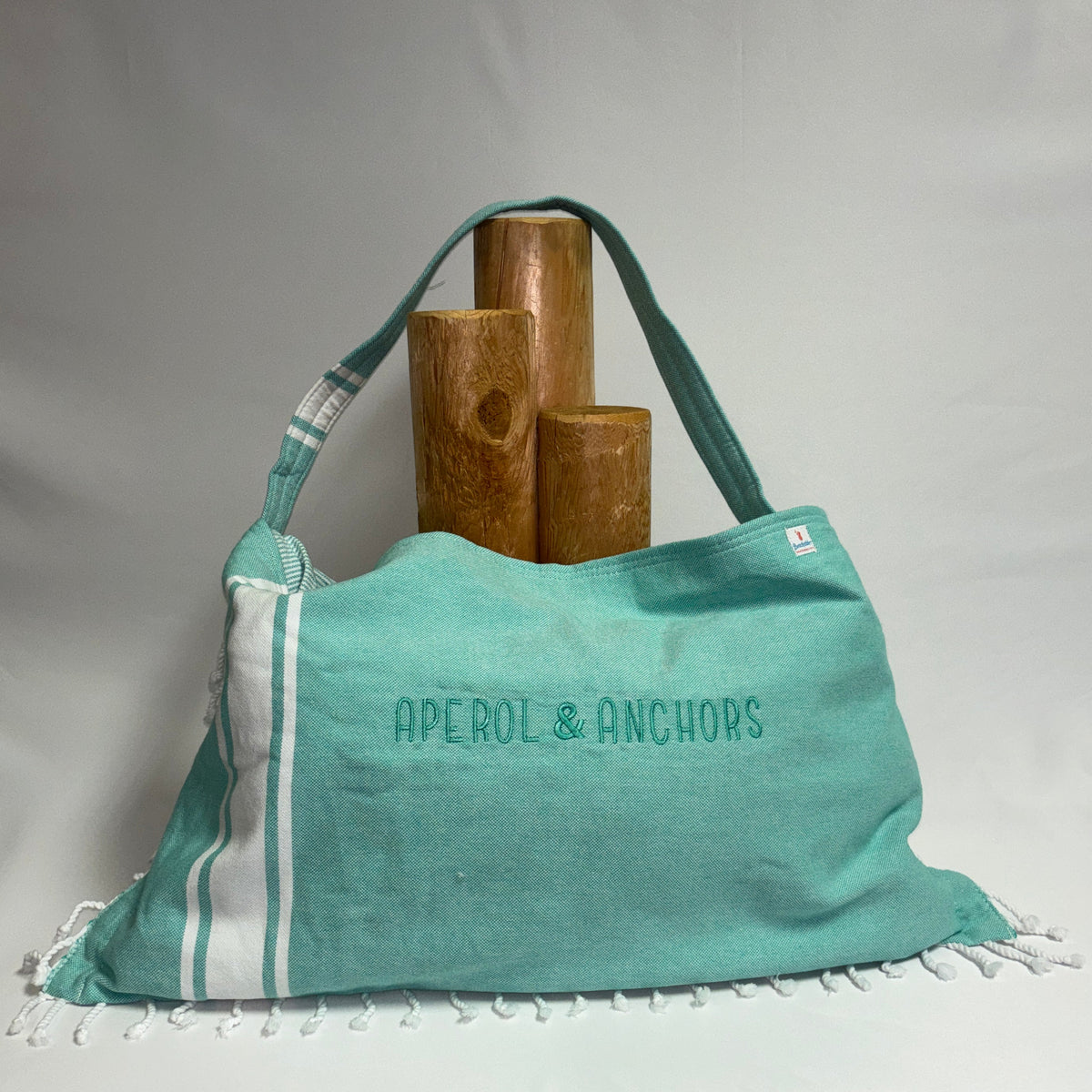 A turquoise and white striped Aperol & Anchors tote bag with fringed edges, displayed on a white draped fabric background with three wooden props behind it.