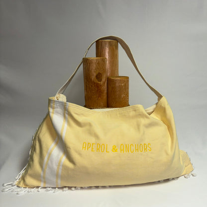 A pale yellow fabric shoulder bag with white stripes and white fringe, featuring the text 'APEROL & ANCHORS' in yellow on the front. The bag is styled with three wooden cylinder props against a neutral gray studio background.