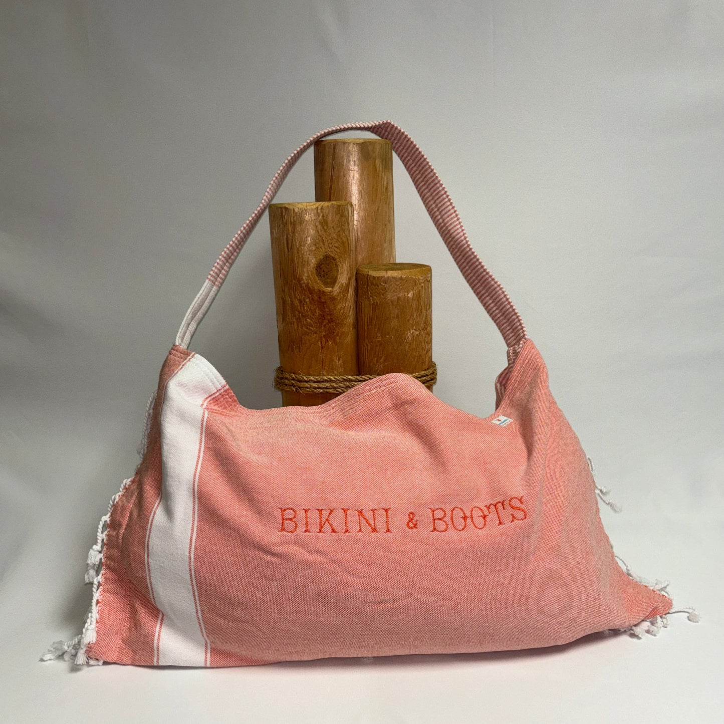 A coral pink and white striped fabric shoulder bag with 'BIKINI & BOOTS' embroidered in coral pink on the front, displayed against a light gray background with decorative wooden props.