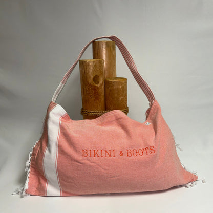 A coral pink and white striped fabric shoulder bag with 'BIKINI & BOOTS' embroidered in coral pink on the front, displayed against a light gray background with decorative wooden props.