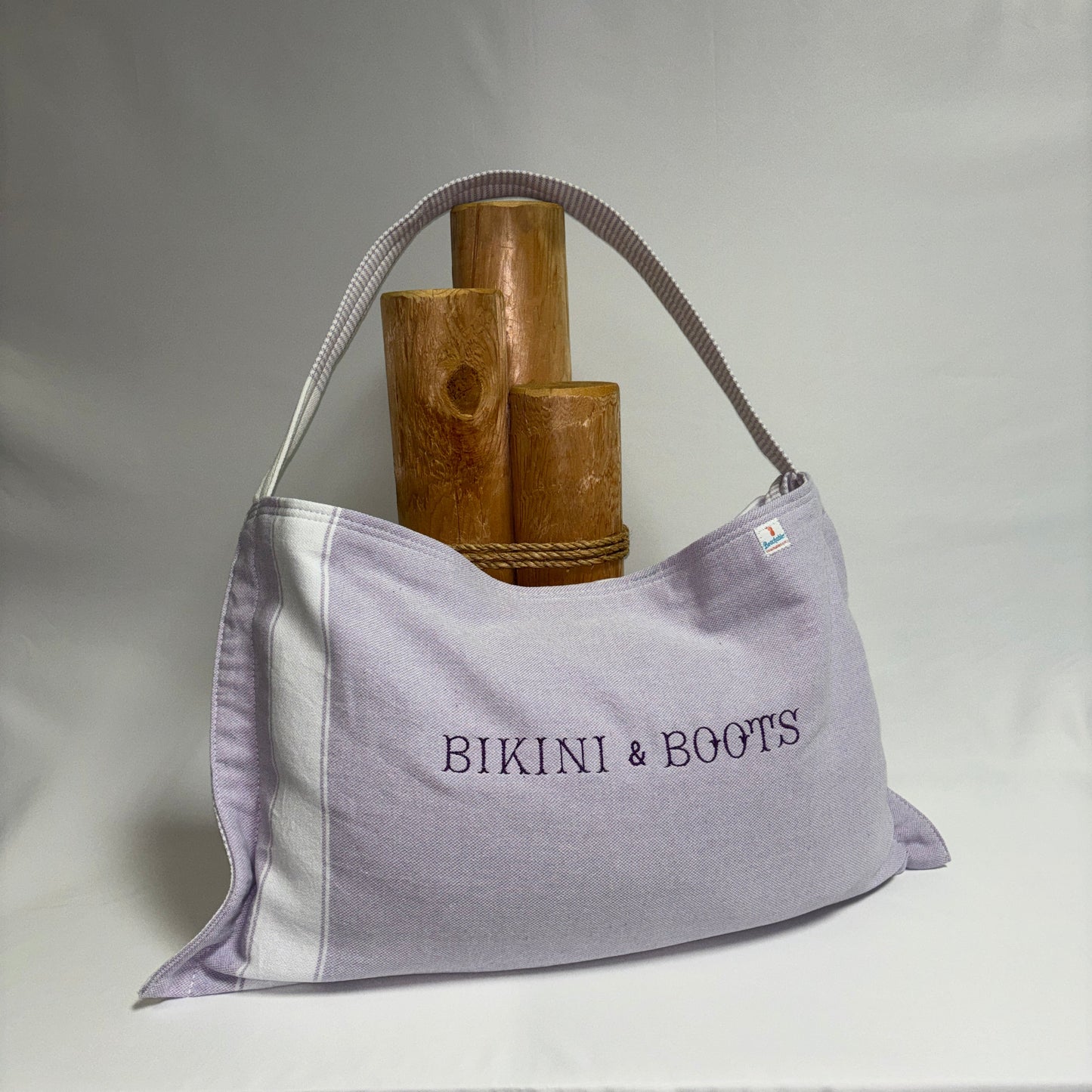 A light lavender tote bag with white side panels and a gray strap, featuring the text 'BIKINI & BOOTS' printed on the front. The bag is styled with three wooden props on a draped white fabric background.