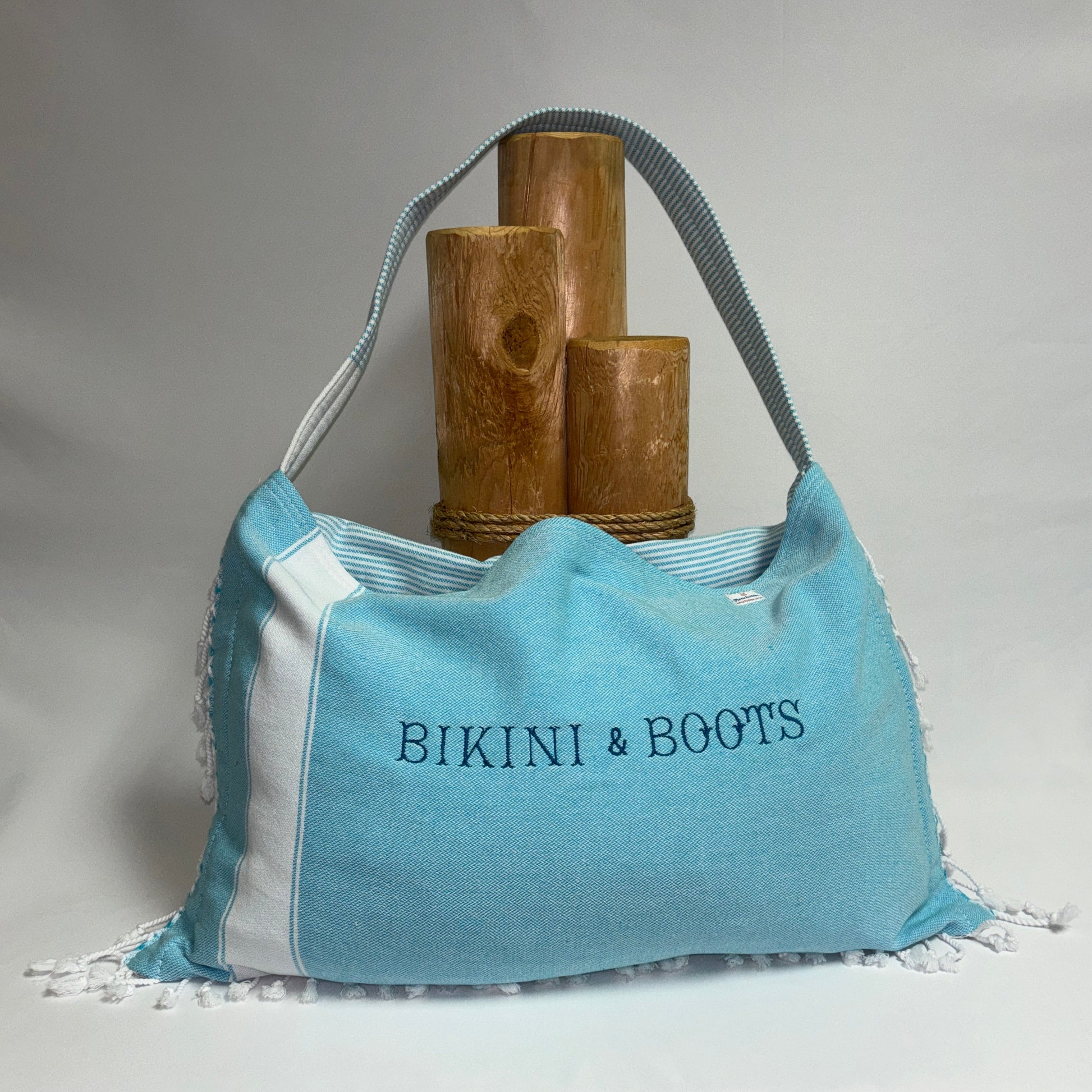 A light blue and white fringed tote bag with the text 'BIKINI & BOOTS' printed on the front, displayed on a white draped surface with three wooden props behind it.