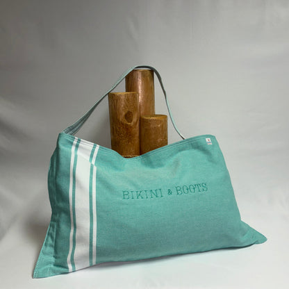 A teal canvas tote bag with white stripes and 'BIKINI & BOOTS' printed on the front, displayed on a draped white fabric backdrop with wooden props behind it.