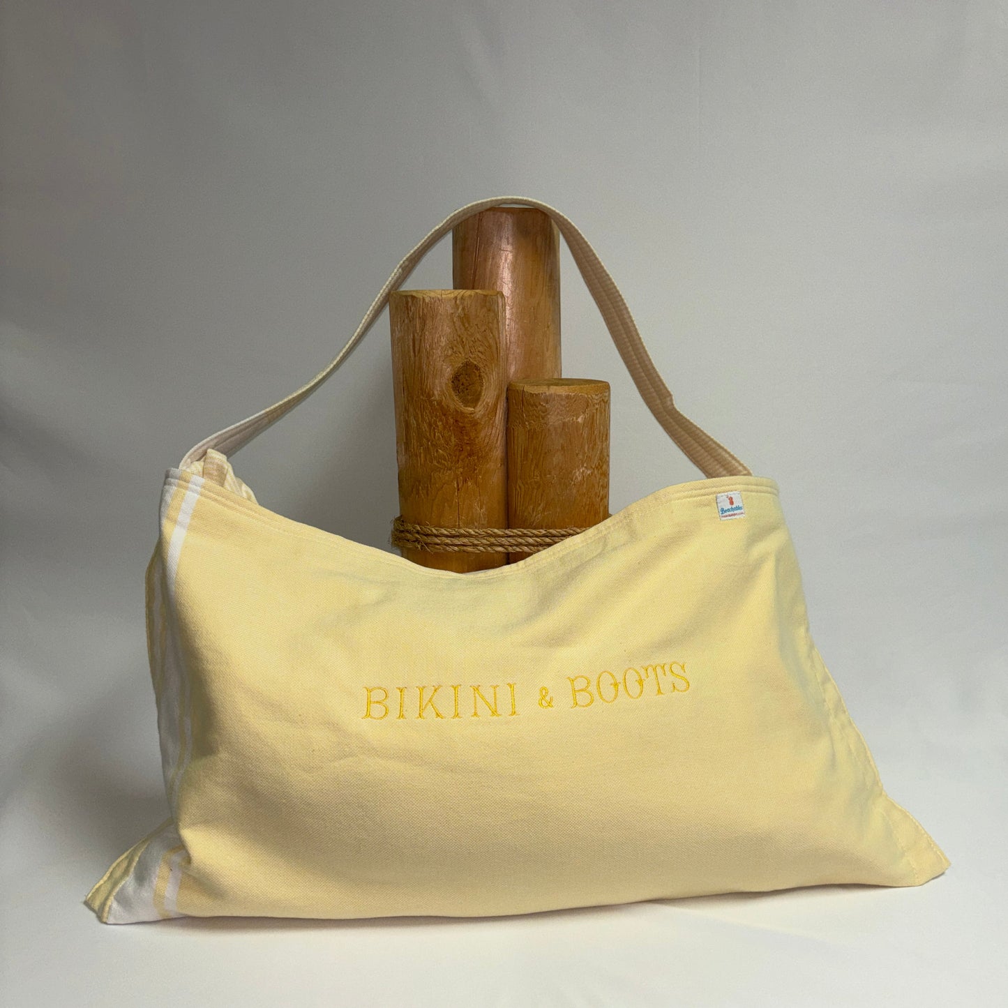 A yellow canvas shoulder bag with yellow embroidered text reading 'BIKINI & BOOTS' sits on a draped white fabric surface with a wooden stand behind it.