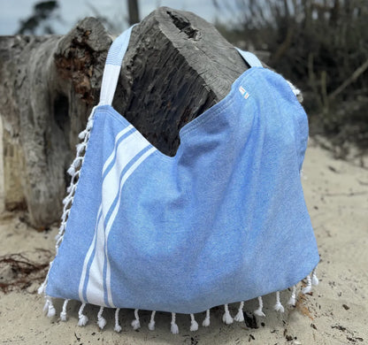 Blue and white beach towel with tassels draped over a log on a sandy beach.