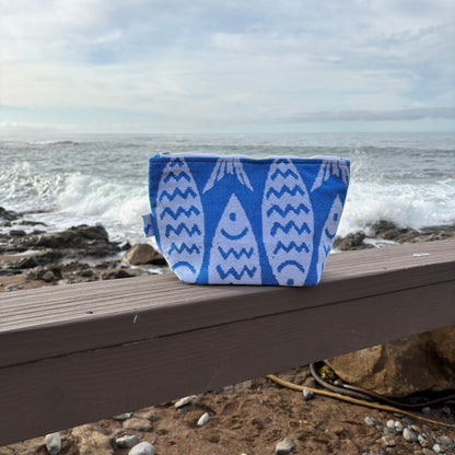 large blue and white fish print makeup bags placed on a wooden bench with a coastal background.