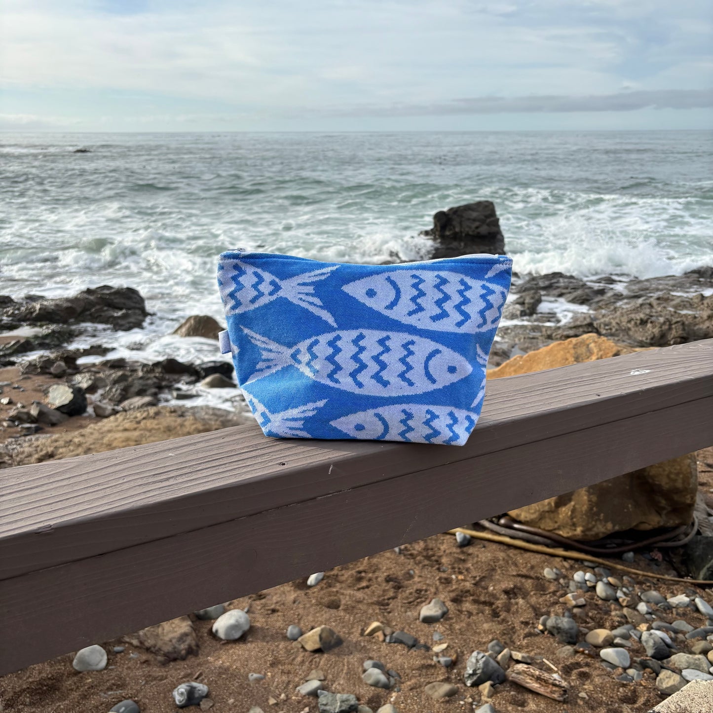 medium sized blue and white fish print makeup bags placed on a wooden bench with a coastal background.