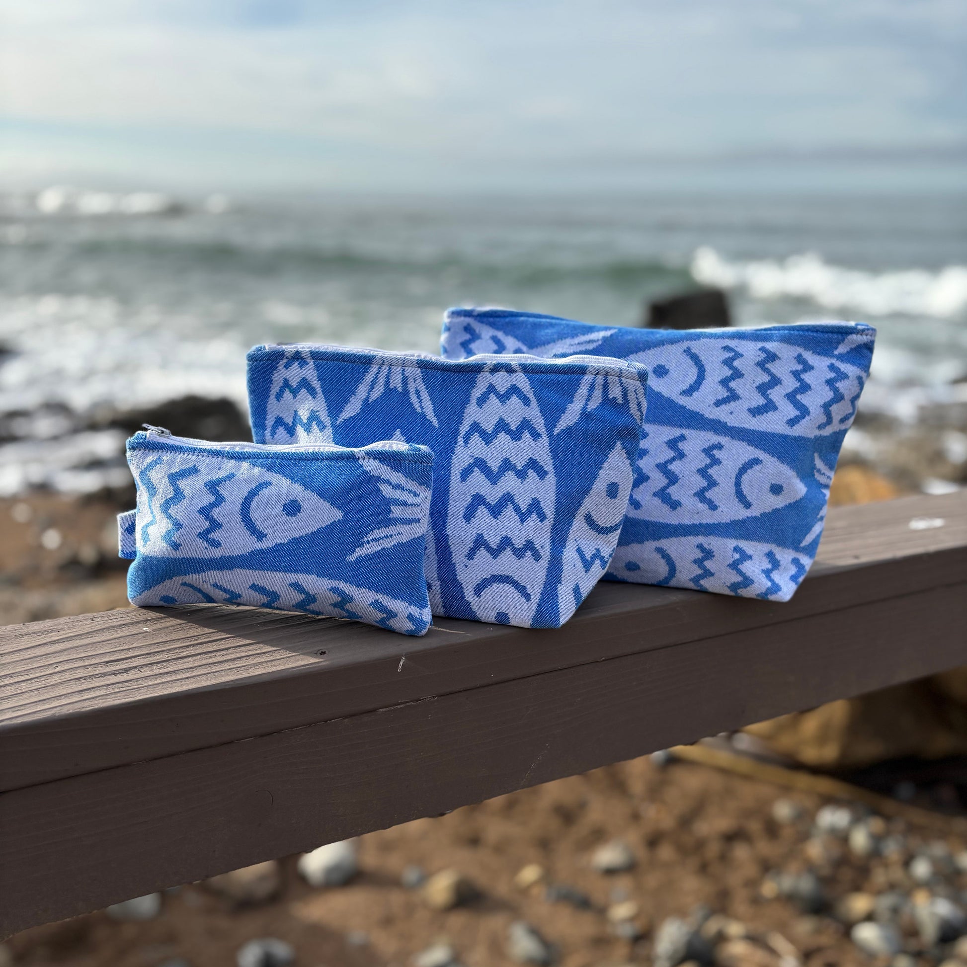 Three blue makeup bags with white fish patterns placed on a wooden bench with a coastal background.