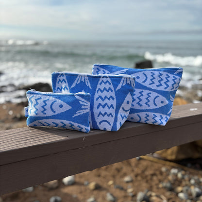 Three blue makeup bags with white fish patterns placed on a wooden bench with a coastal background.