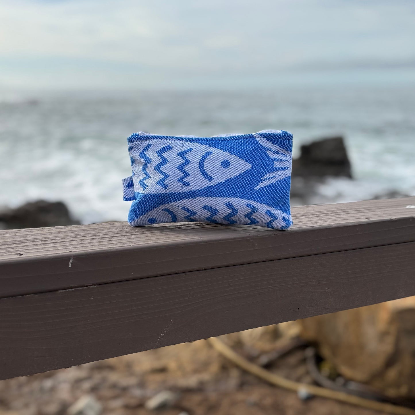 small blue and white fish print makeup bags placed on a wooden bench with a coastal background.