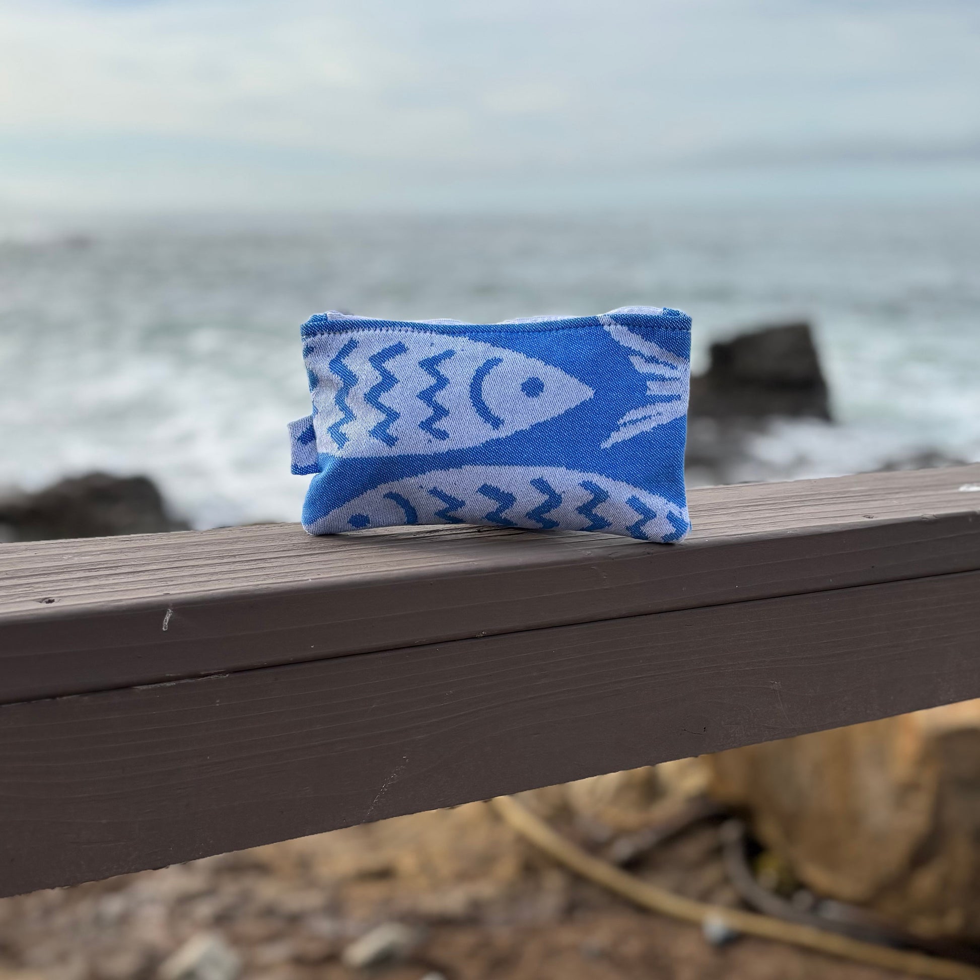 small blue and white fish print makeup bags placed on a wooden bench with a coastal background.