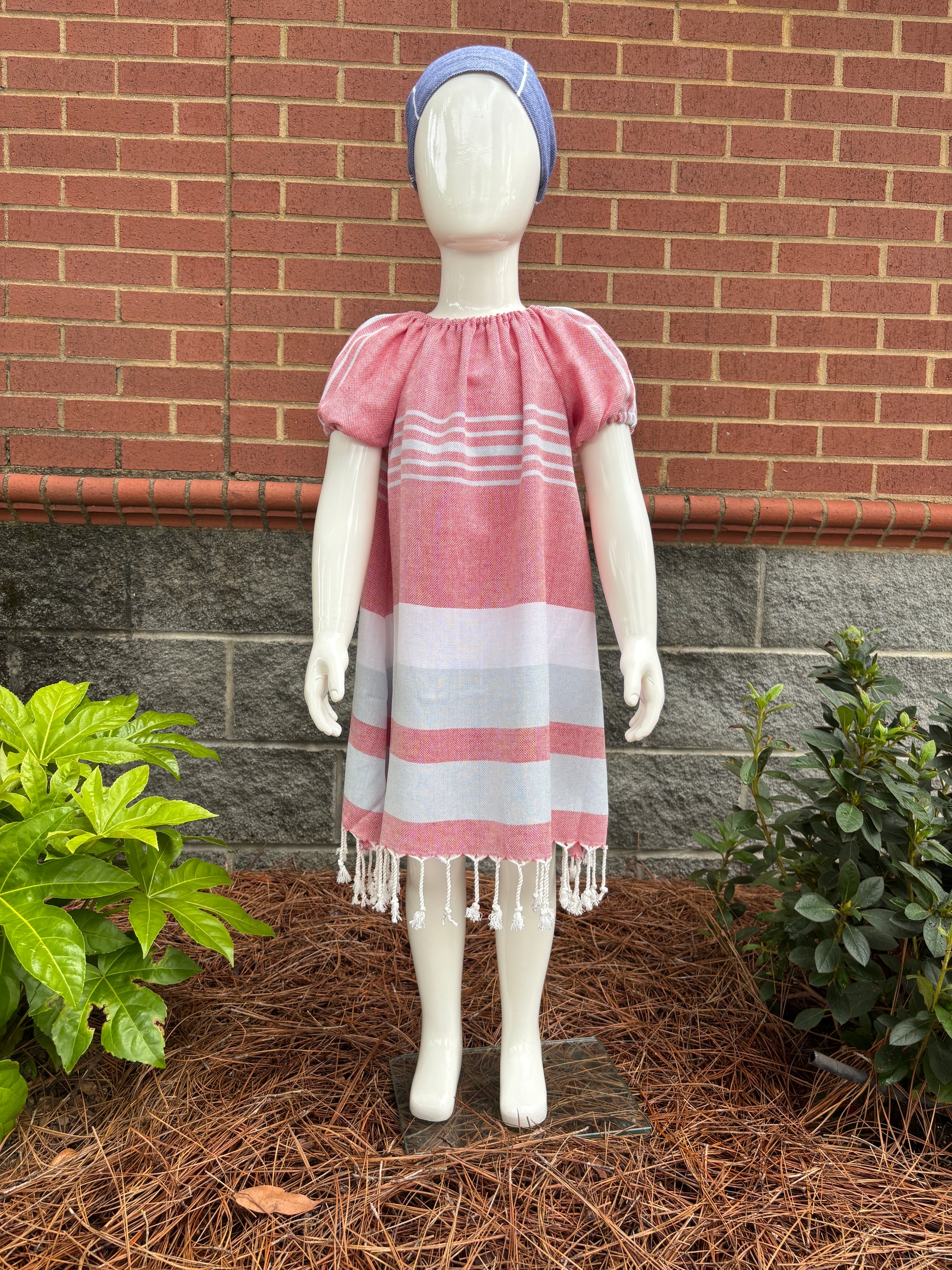 Mannequin wearing  blue red and white striped dress with puff sleeves and tassels at hem in front of red brick wall