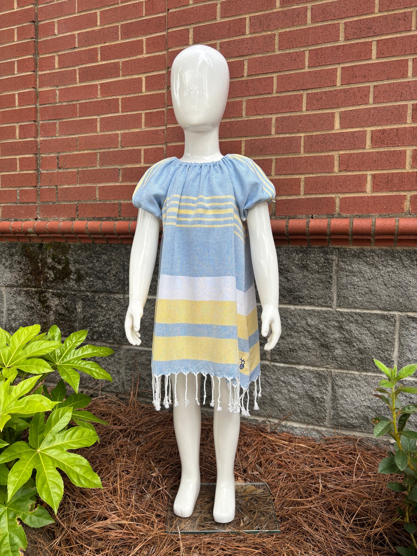 Mannequin wearing blue, yellow and white striped dress with puff sleeves and tassels at hem in front of red brick wall