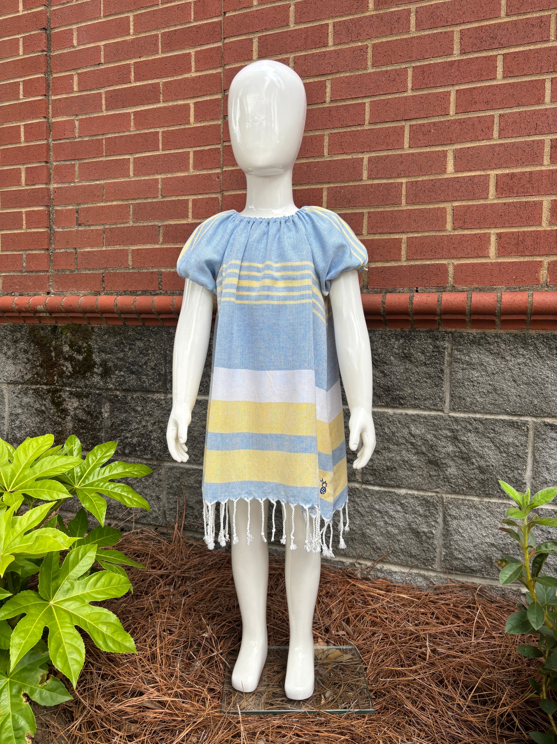 Mannequin wearing blue, yellow and white striped dress with puff sleeves and tassels at hem in front of red brick wall