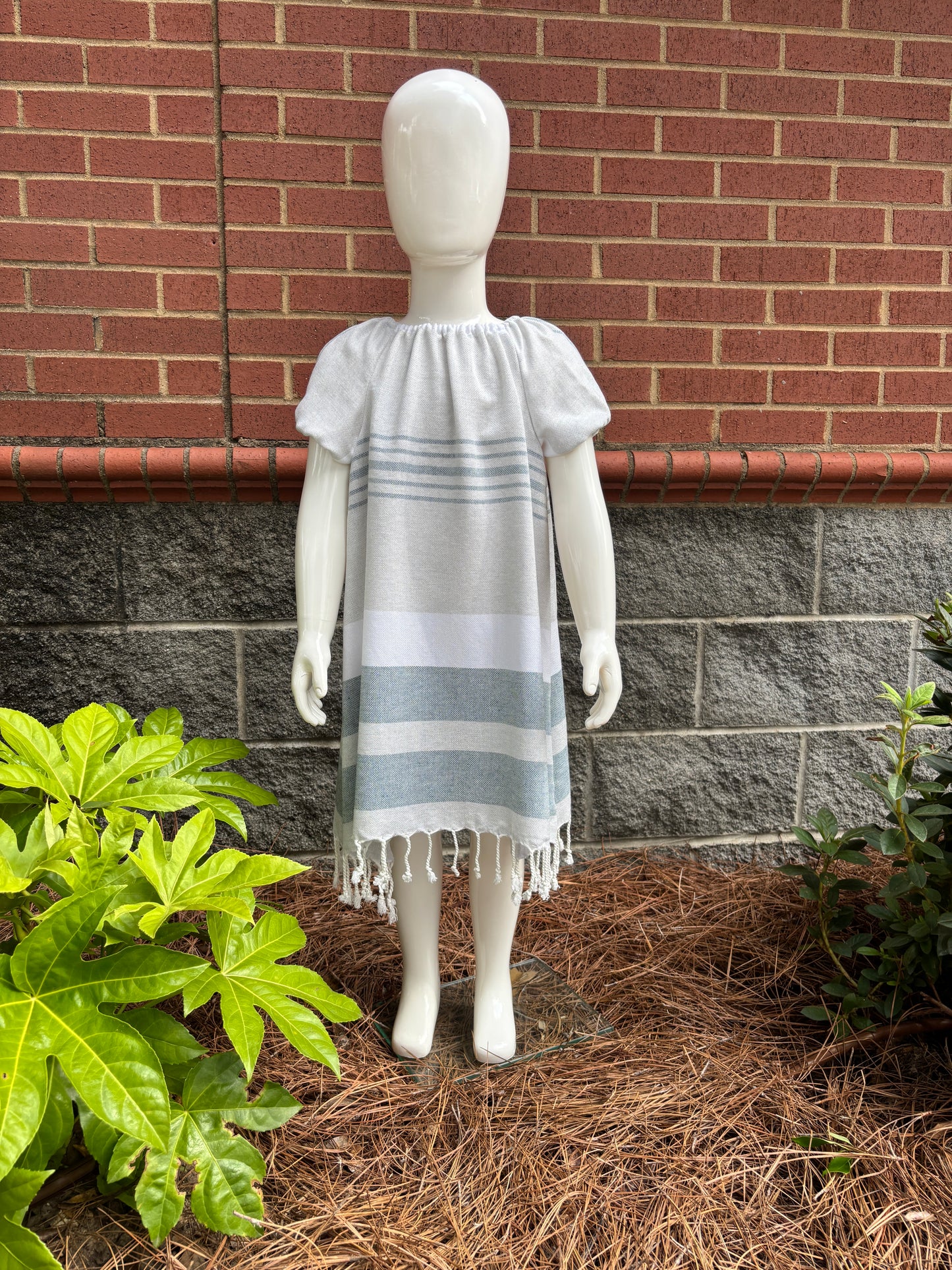 Mannequin wearing  charcoal, grey and white striped dress with puff sleeves and tassels at hem in front of red brick wall