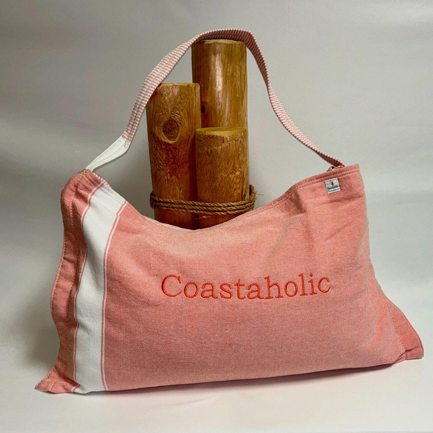 A coral and white striped canvas shoulder bag with 'Coastaholic' embroidered in orange on the front, styled with three wooden log props on a neutral light gray background.