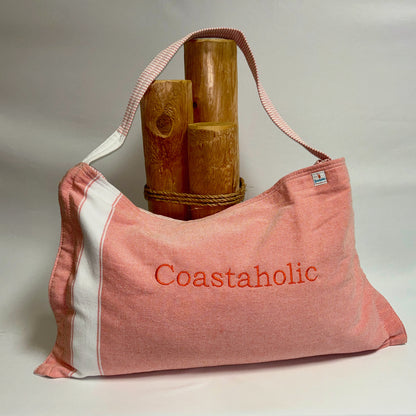 A coral and white striped canvas shoulder bag with 'Coastaholic' embroidered in orange on the front, styled with three wooden log props on a neutral light gray background.