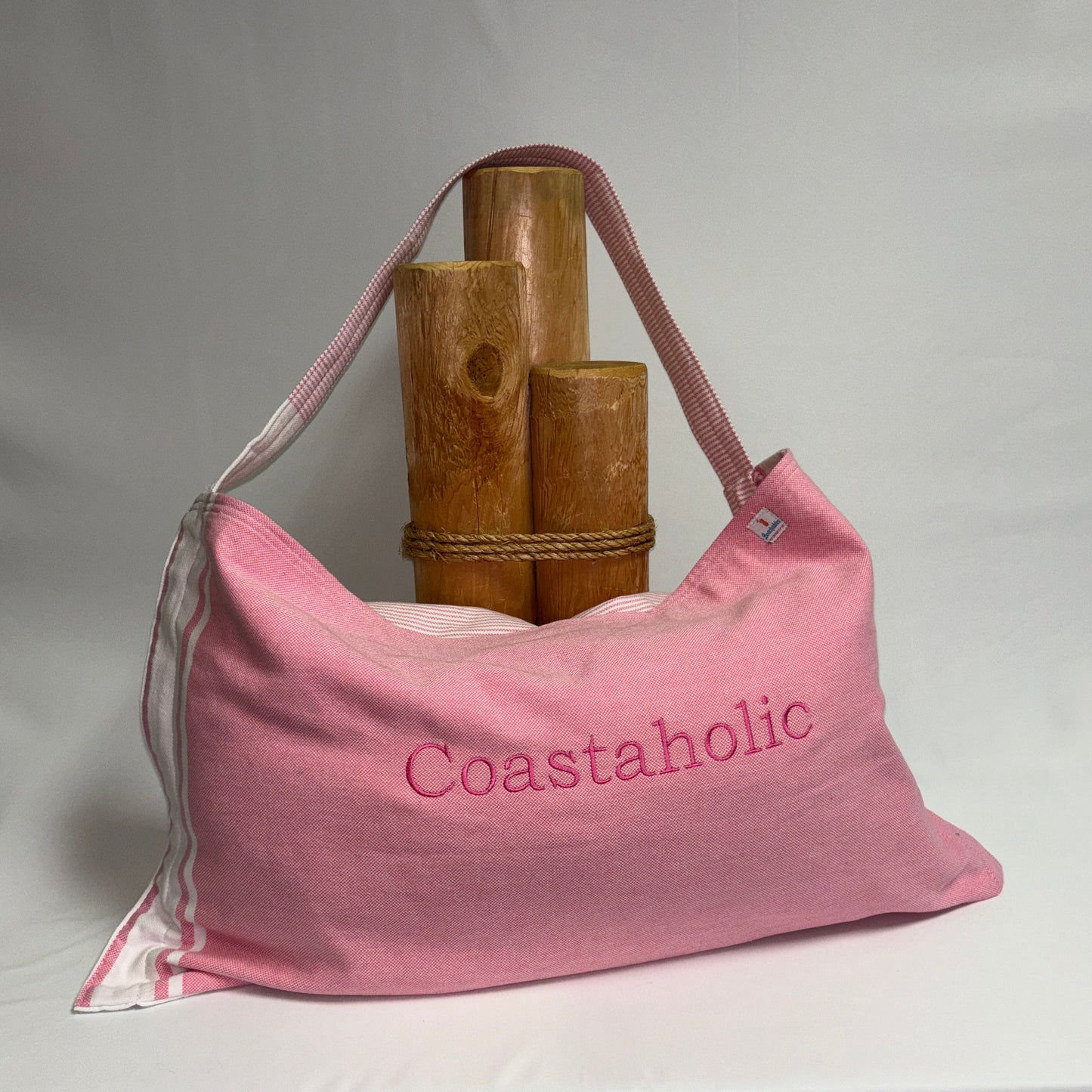 A pink canvas shoulder bag with 'Coastaholic' embroidered on the front, displayed on a draped white fabric surface with a rustic bamboo stand behind it.