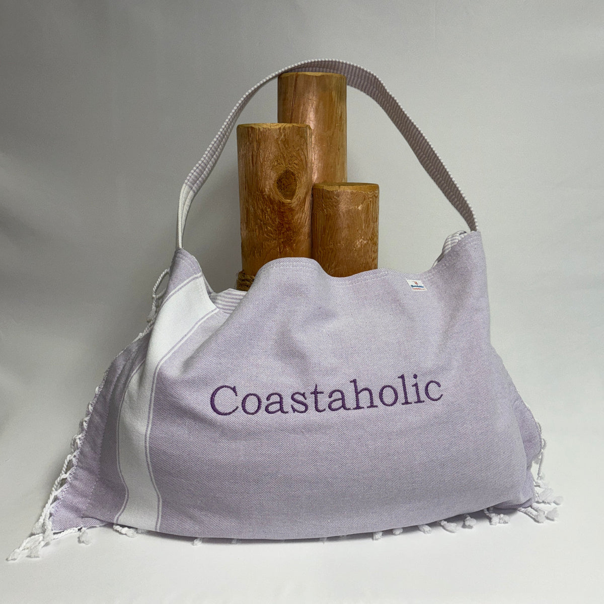 A large light purple and white slouchy shoulder bag with 'Coastaholic' embroidered on the front, styled with three wooden cylinder props against a draped white fabric background.