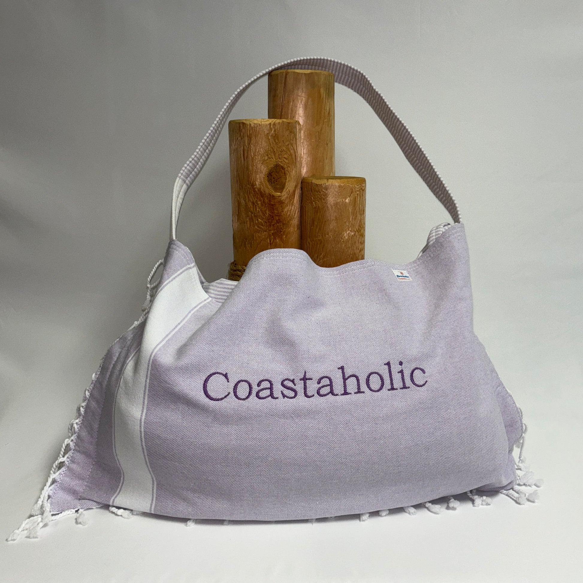 A large light purple and white slouchy shoulder bag with 'Coastaholic' embroidered on the front, styled with three wooden cylinder props against a draped white fabric background.