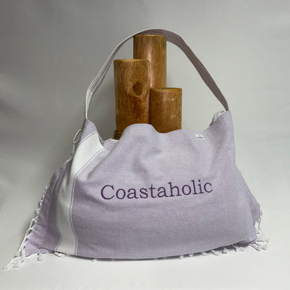 A large light purple and white slouchy shoulder bag with 'Coastaholic' embroidered on the front, styled with three wooden cylinder props against a draped white fabric background.