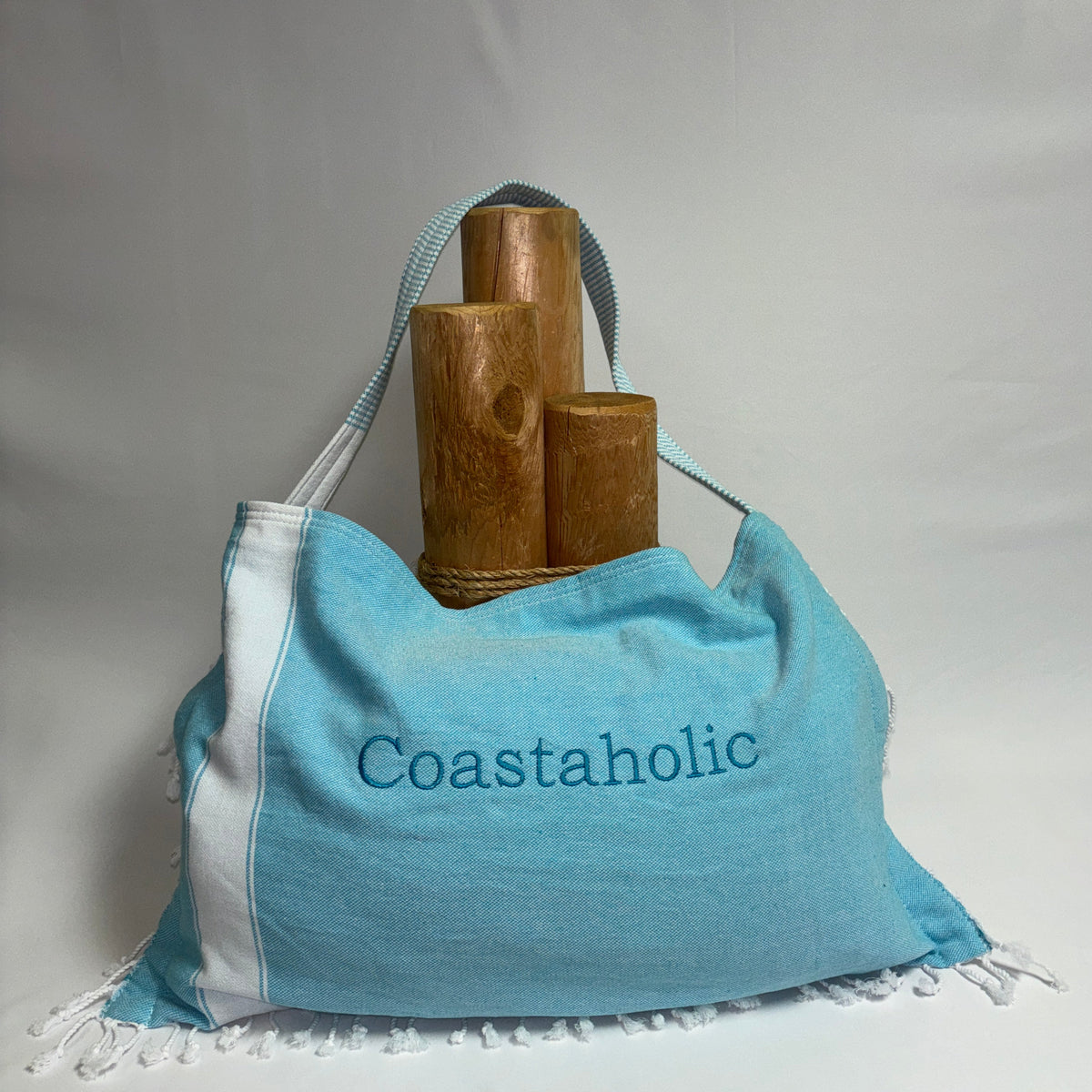 A light blue fabric tote bag with white side panels and white fringe, featuring the word 'Coastaholic' printed in blue on the front. The bag is shown holding three wooden cylindrical props against a white fabric backdrop.