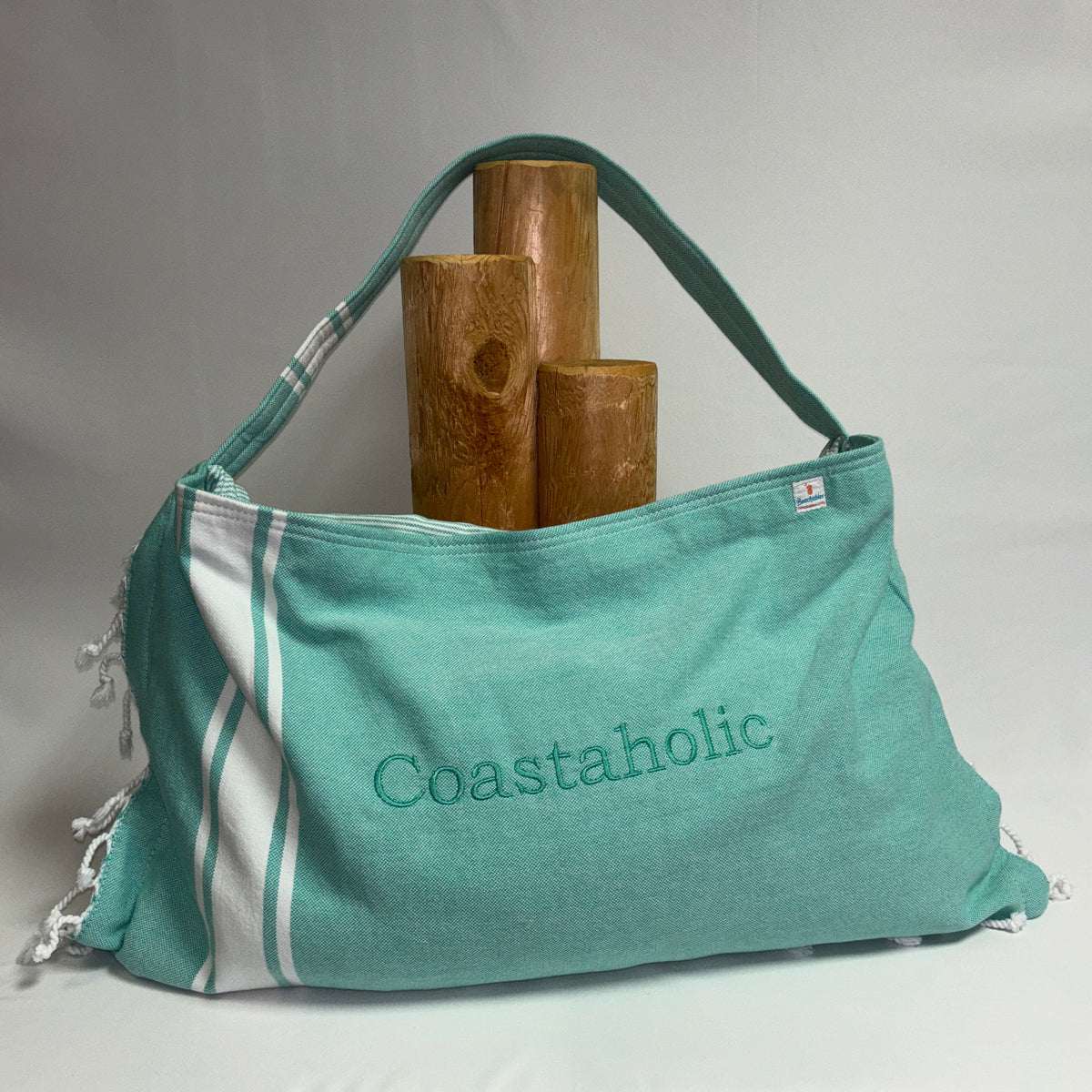 A teal and white Coastaholic canvas shoulder bag with embroidered branding, displayed on a draped white fabric surface with three wooden logs behind it.