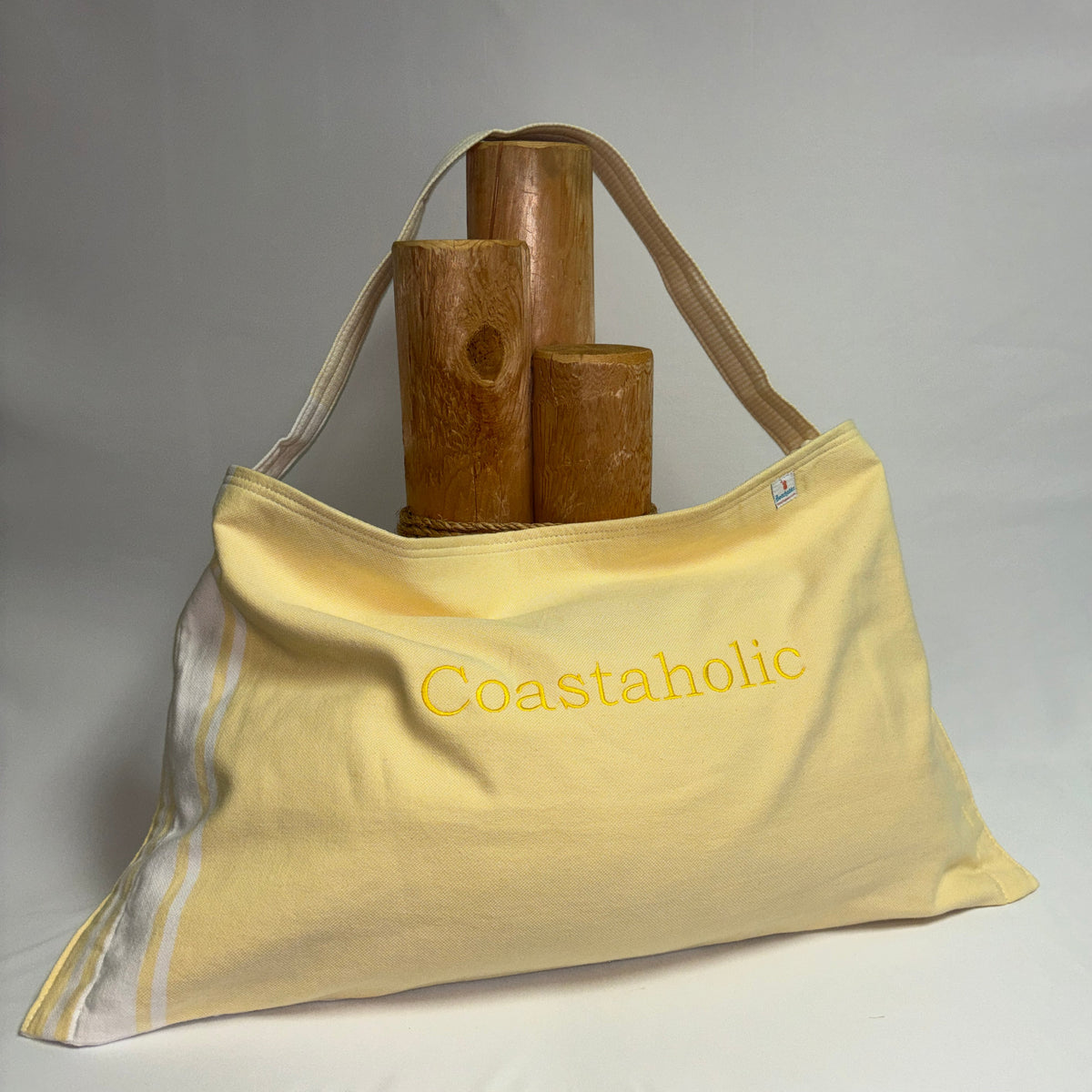 A pale yellow Coastaholic canvas tote bag with a single shoulder strap, featuring the brand name embroidered in yellow on the front. The bag is styled with three wooden cylinder props on top, set against a neutral gray fabric backdrop.