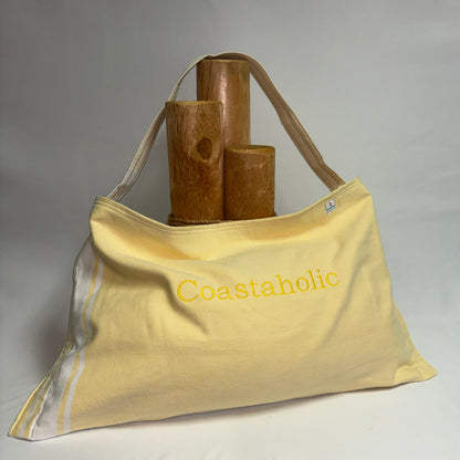 A pale yellow Coastaholic canvas tote bag with a single shoulder strap, featuring the brand name embroidered in yellow on the front. The bag is styled with three wooden cylinder props on top, set against a neutral gray fabric backdrop.