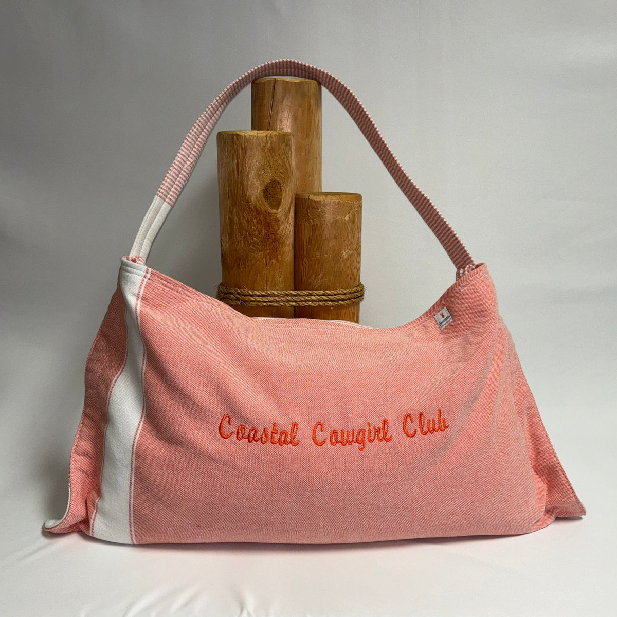 A pink canvas shoulder bag with white side panels and orange embroidered text reading 'Coastal Cowgirl Club,' displayed against a white draped backdrop with wooden props behind it.