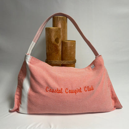 A pink canvas shoulder bag with white side panels and orange embroidered text reading 'Coastal Cowgirl Club,' displayed against a white draped backdrop with wooden props behind it.