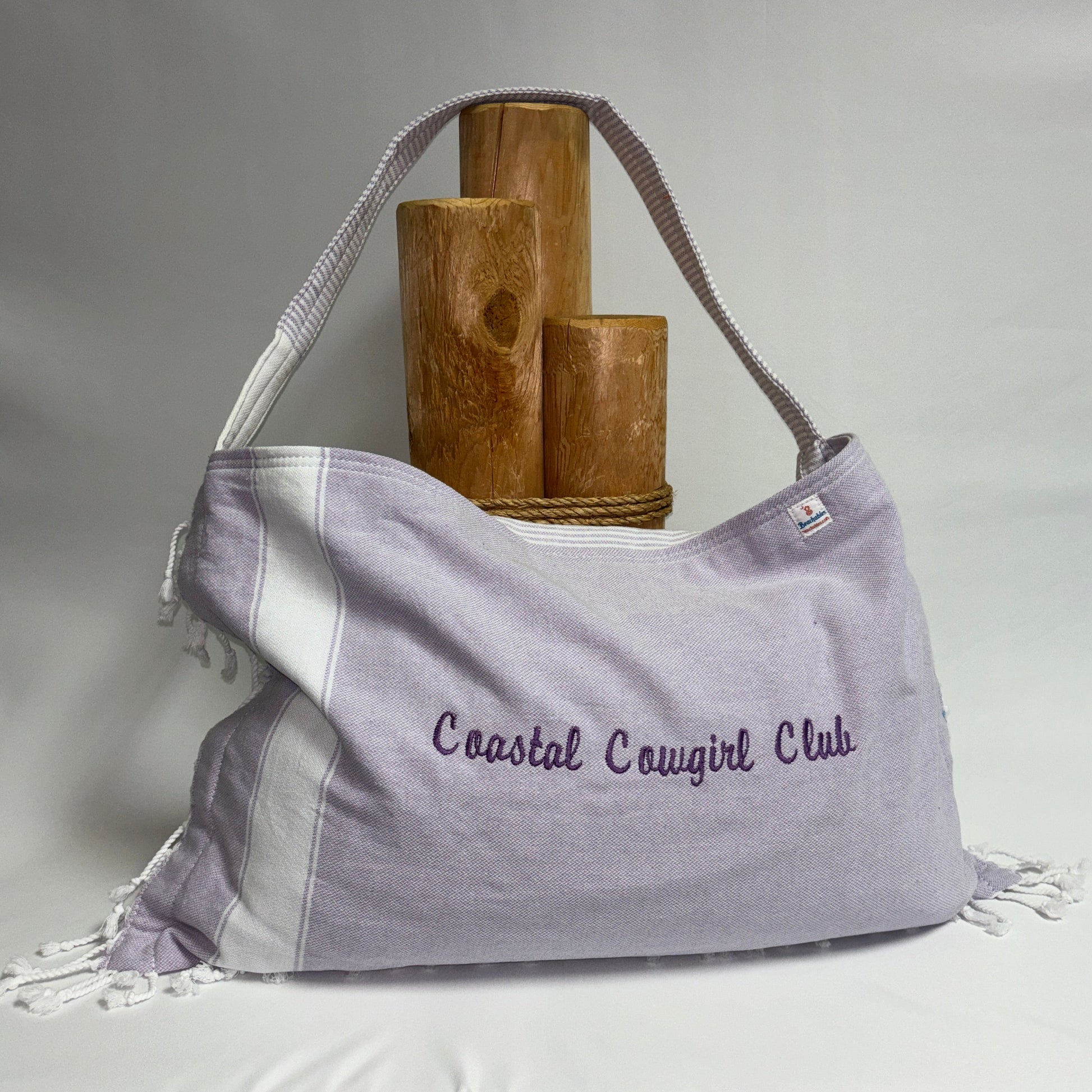 A lavender and white striped fabric shoulder bag with fringe details and the embroidered text 'Coastal Canguirl Club' on the front, displayed on a draped white fabric surface with wooden props behind it.
