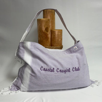 A lavender and white striped fabric shoulder bag with fringe details and the embroidered text 'Coastal Canguirl Club' on the front, displayed on a draped white fabric surface with wooden props behind it.
