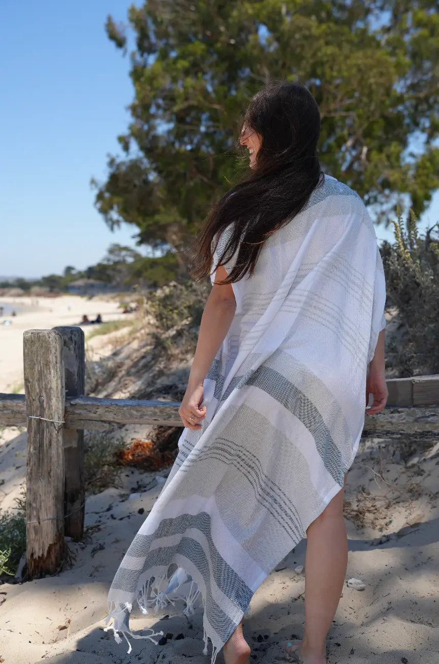 Woman standing on a sandy beach wearing a white and gray striped kimono wrap.
