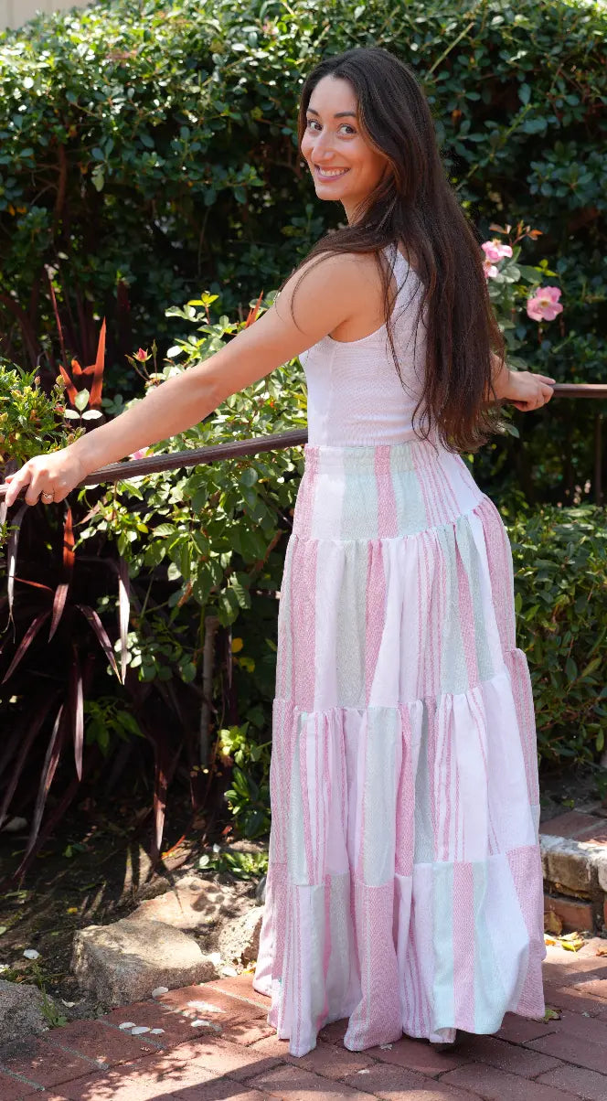 Side view of a Woman wearing a maxi length tiered skirt with pink, green and white stripes with a white tank top outside 