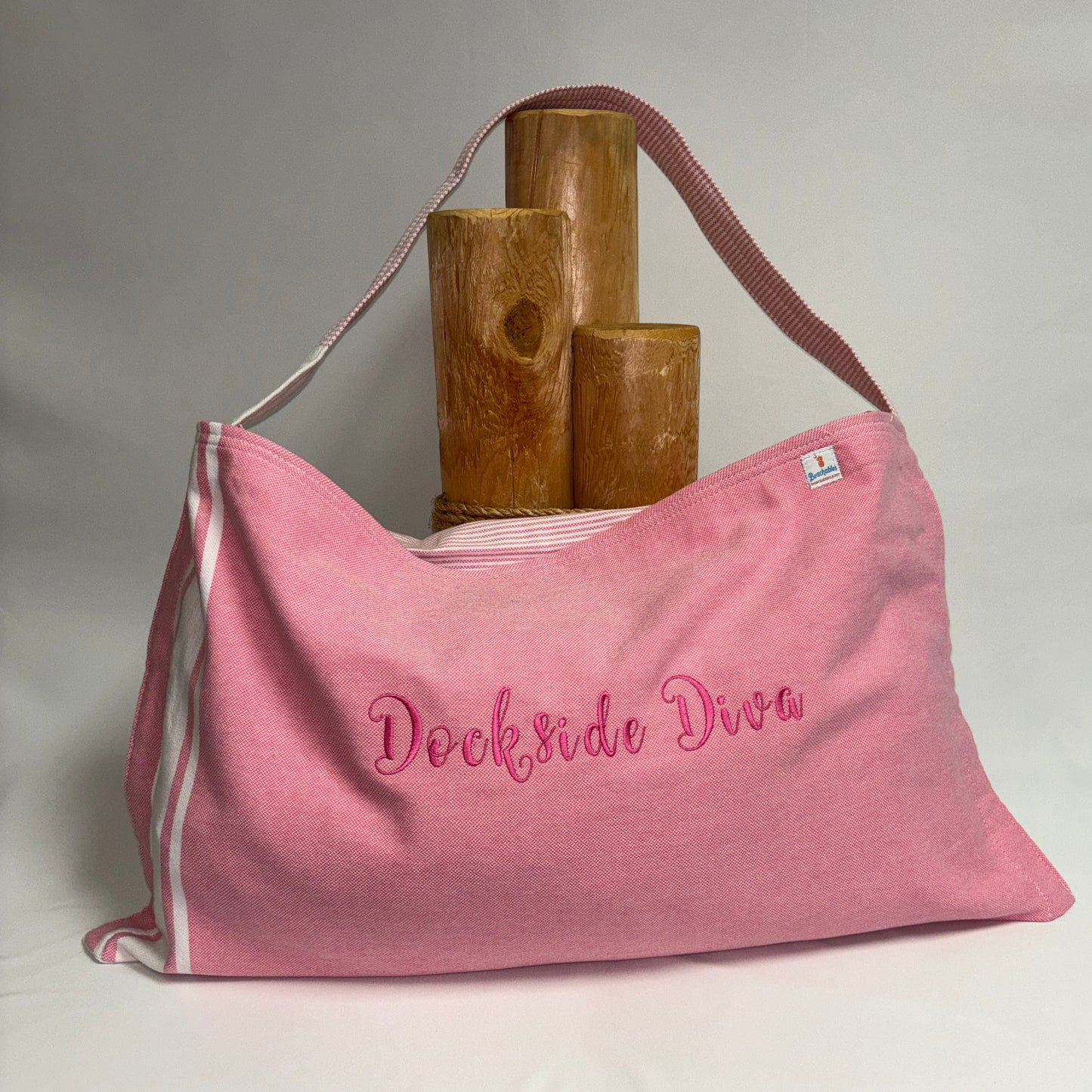 A pink canvas shoulder bag with 'Dockrdo Dream' embroidered in pink script on the front, featuring white side stripes and a pink strap, displayed on a white draped surface with wooden props.