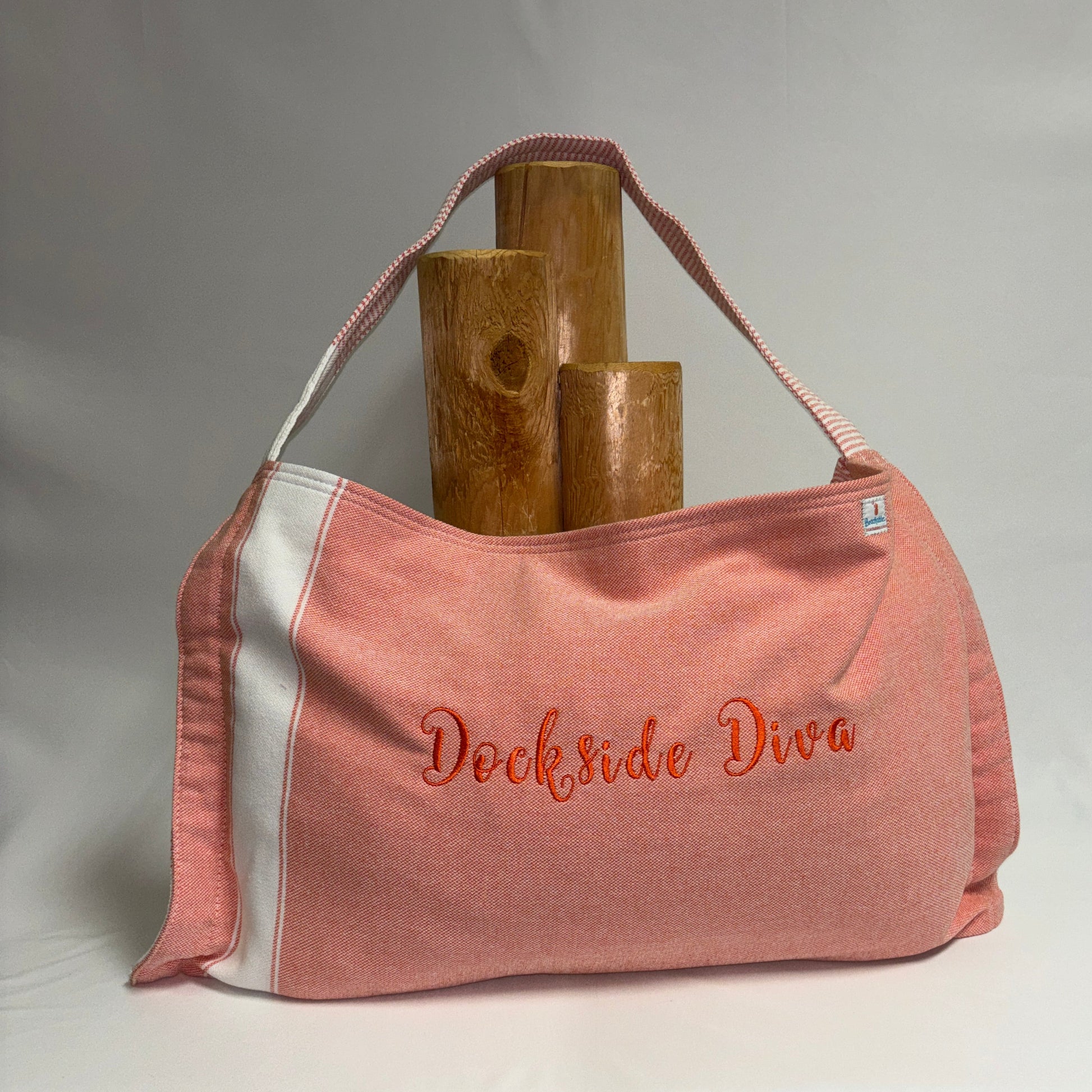 A dusty pink towel bag with white side panels and orange embroidered text reading 'Dockside Diva' on the front, displayed on a white draped surface with three wooden props behind it.
