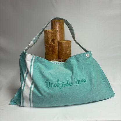 A mint green canvas shoulder bag with white side stripes and 'Dockside Dive' embroidered in teal on the front, displayed on a white draped backdrop with three wooden props.